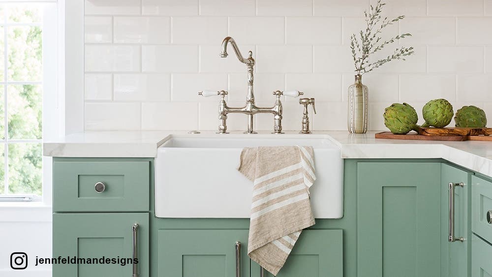 White Farmhouse kitchen sink on green cabinets with light brown towel on the sink.